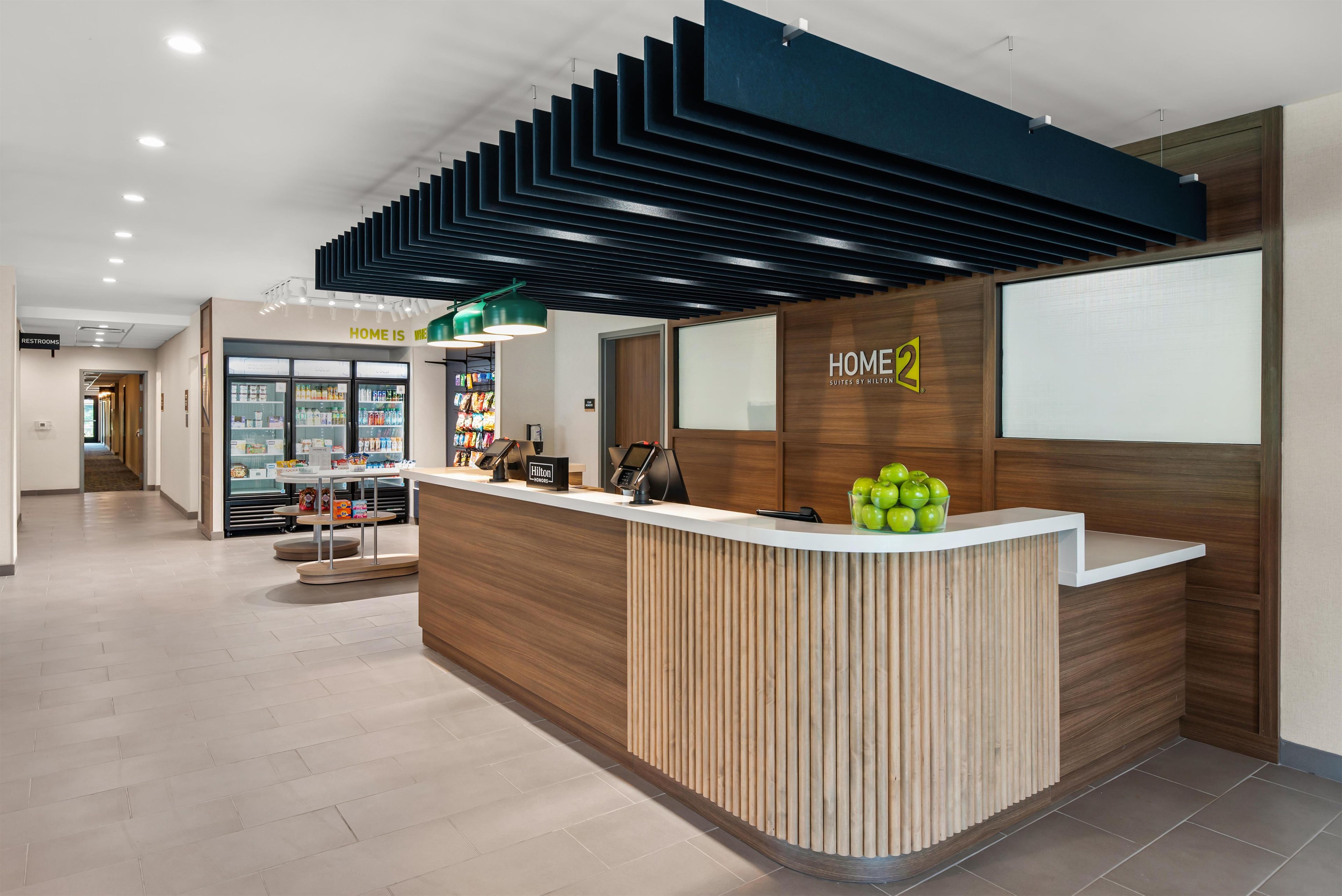 Un mostrador de recepción de hotel con diseño moderno, un cartel que indica 'HOME 2 Suites by Hilton' y un refrigerador con bebidas en el fondo.