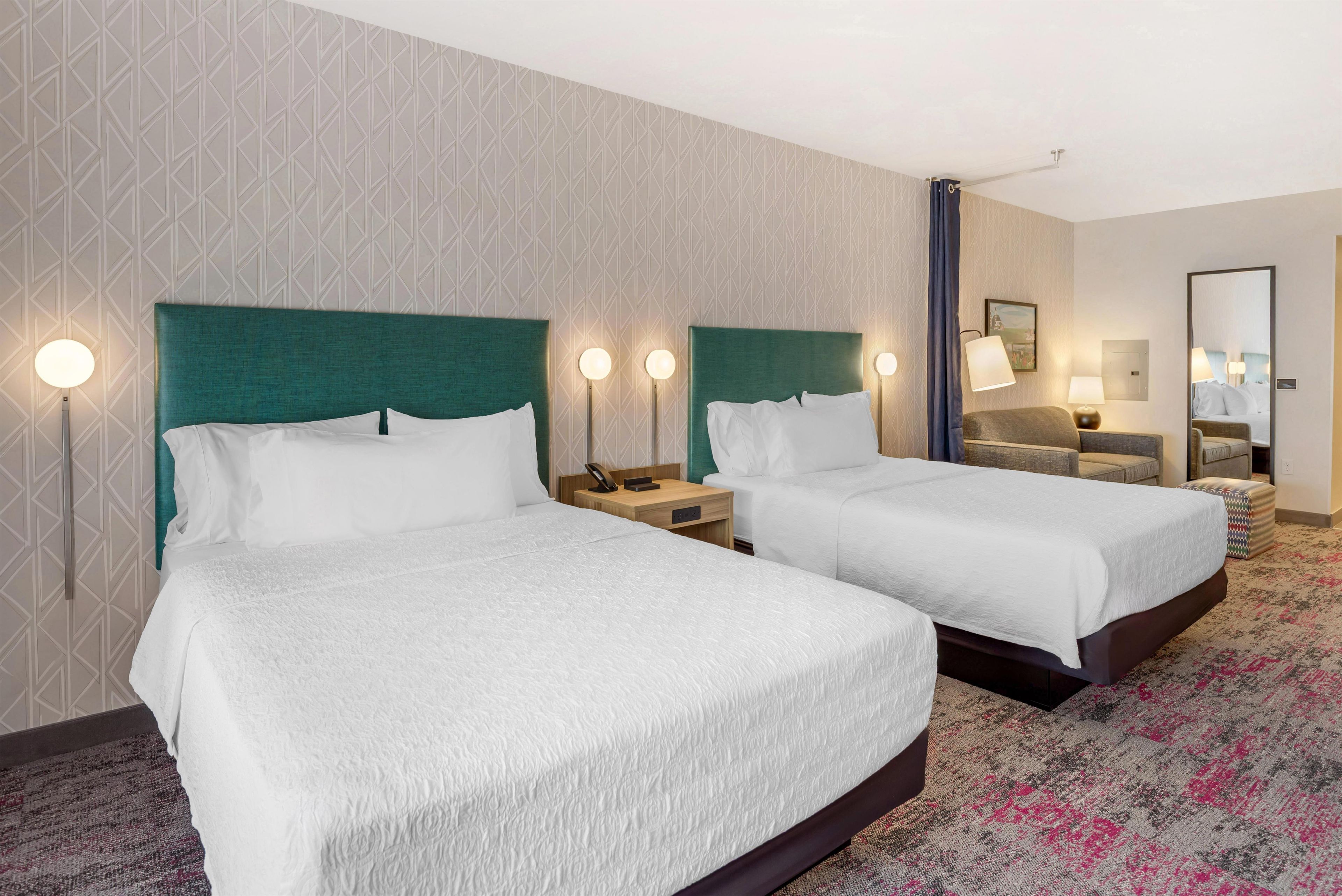 A hotel room with two beds, a nightstand with a lamp, a bedside table with a lamp, and a mirror on the wall.