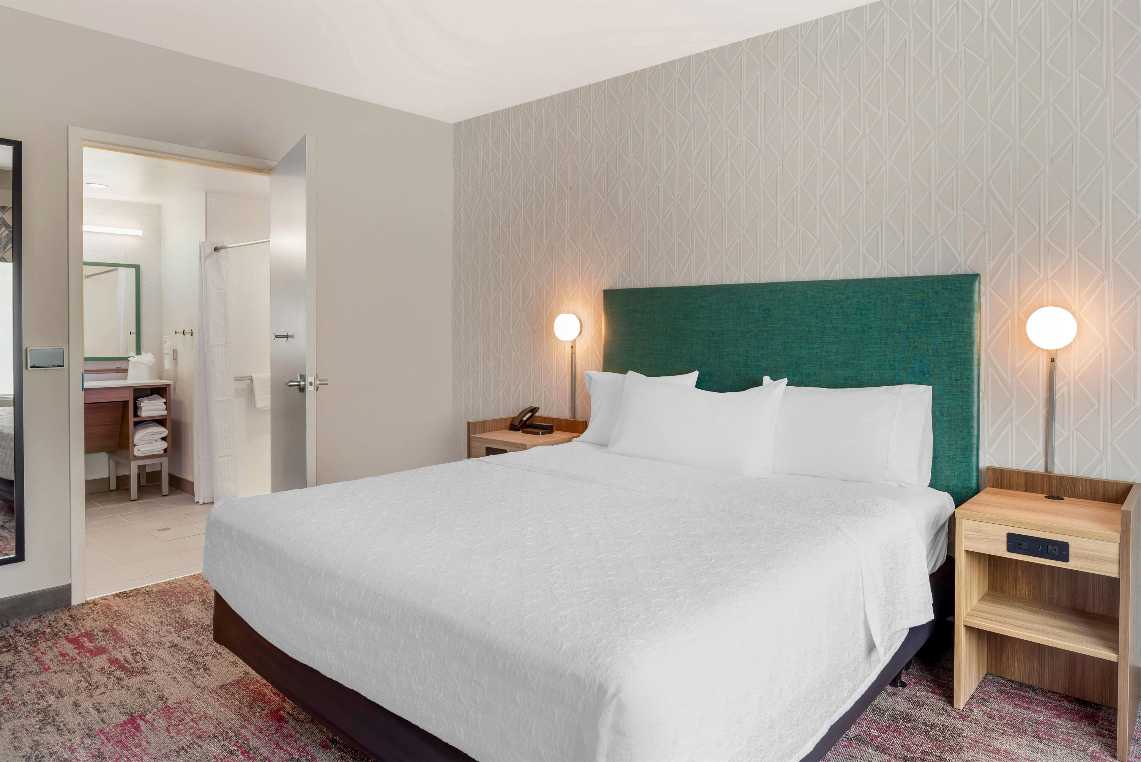 A hotel room with a large bed, two bedside tables, a nightstand, a mirror, a bathroom with a shower, and a patterned wall.