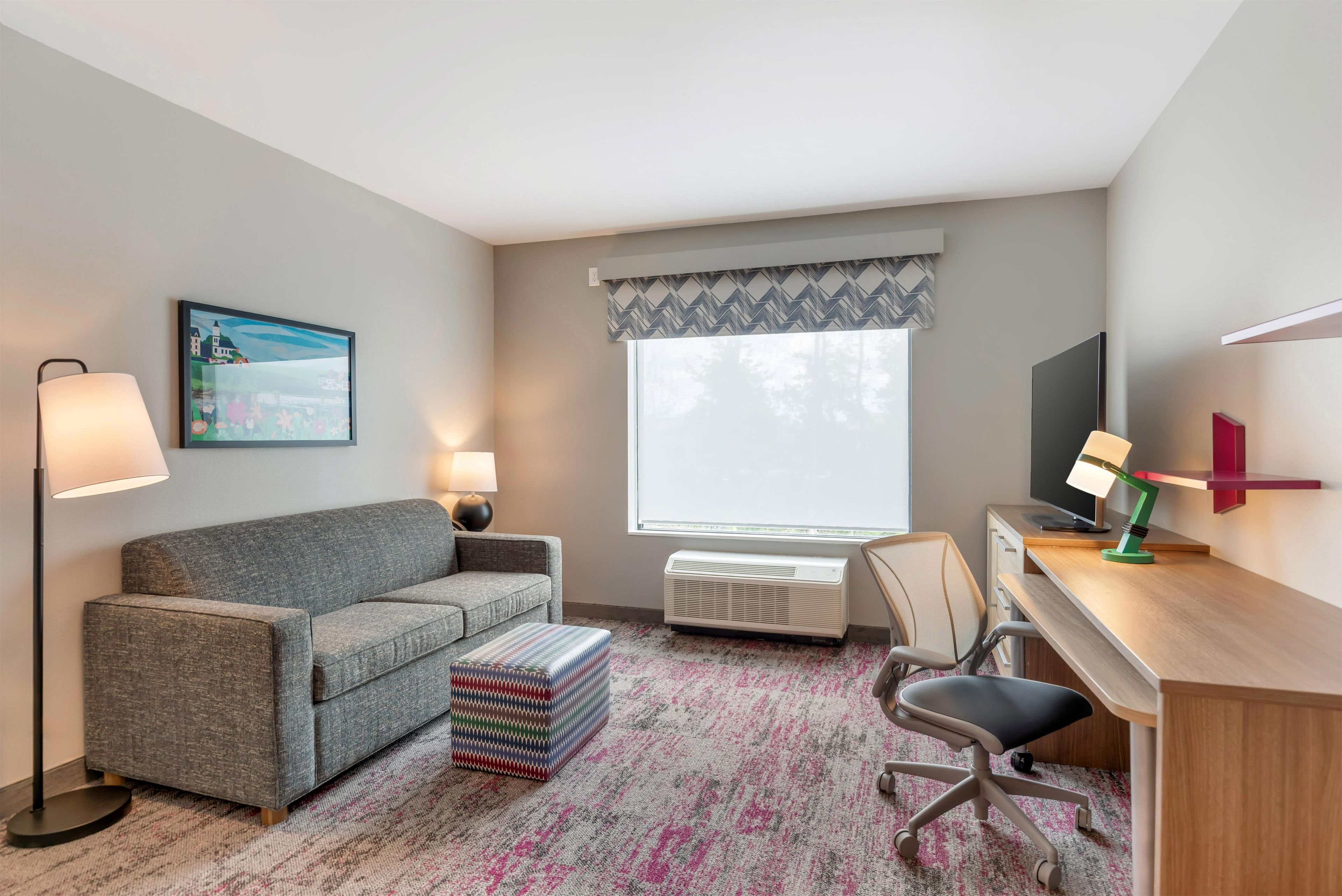 A hotel room with a grey sofa, a small ottoman, a desk with a chair, a television, and a window with a view of trees.