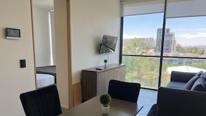 A modern living room with a dining table, a sofa, a TV mounted on the wall, and a large window with a city view. - Livify (Zapopan)