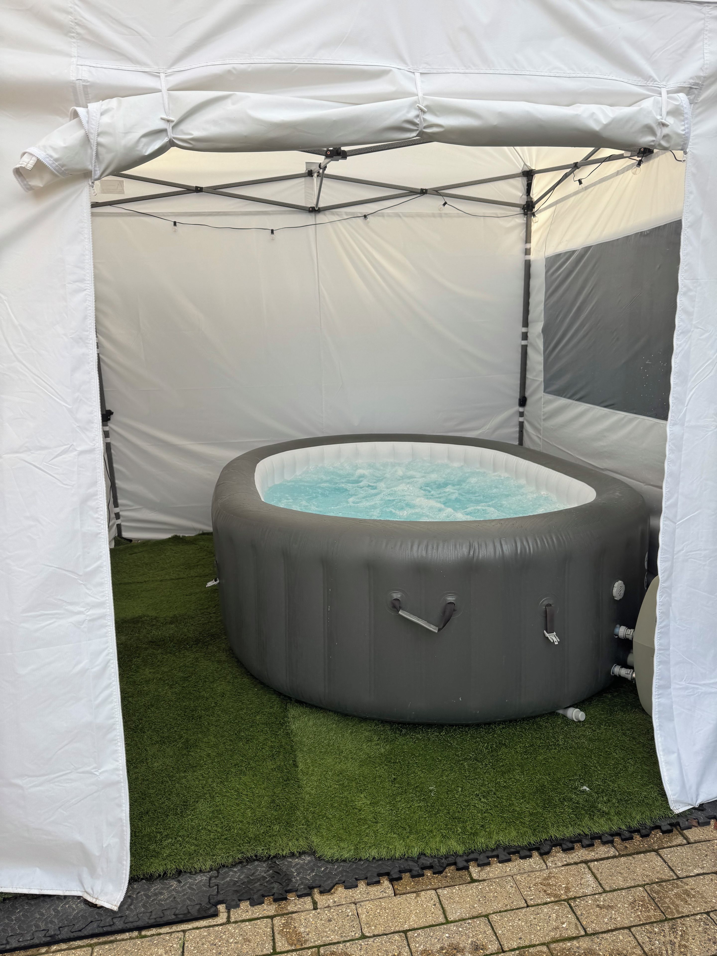 A hot tub under a white canopy with a grassy surface and a brick border.