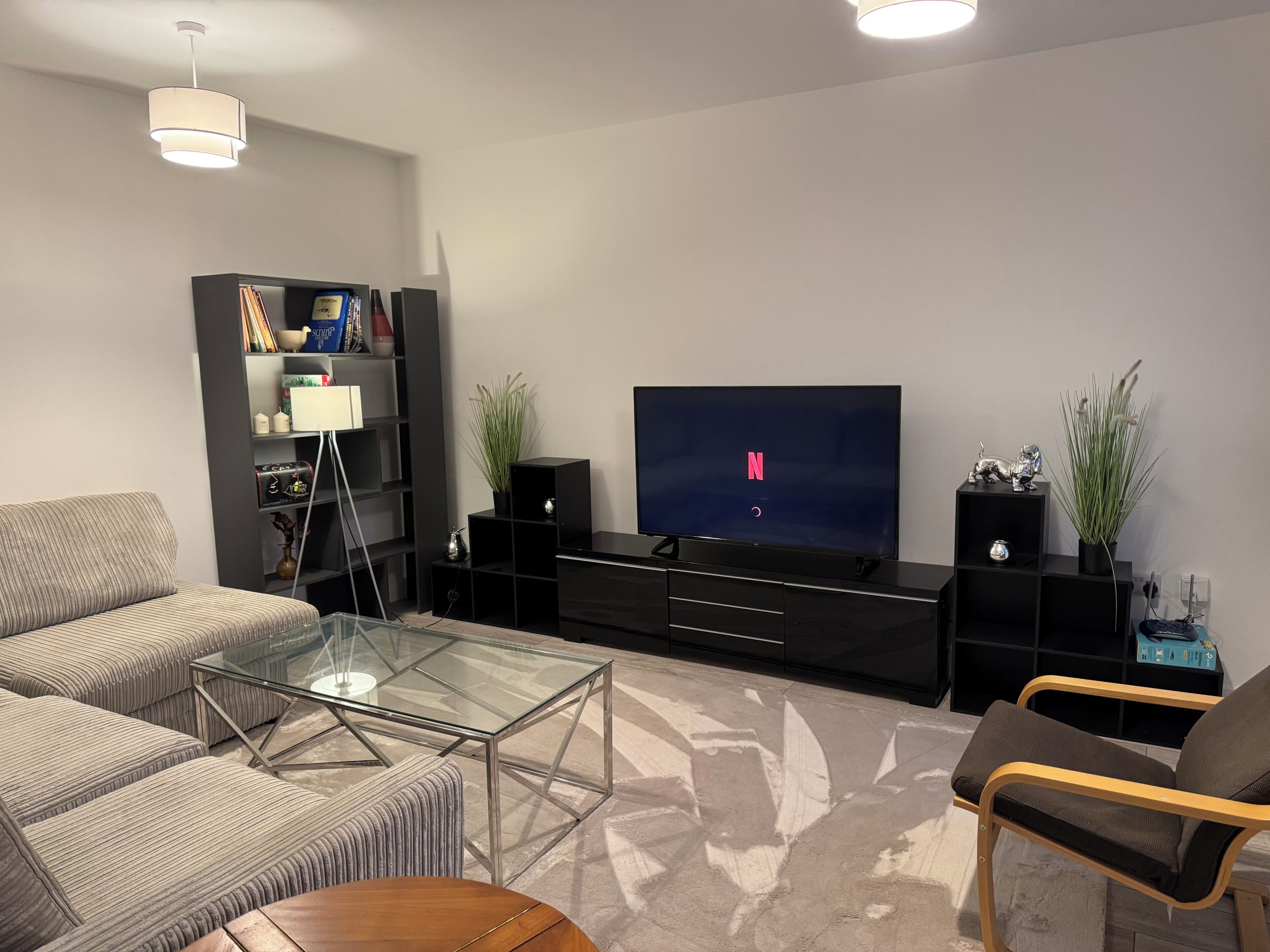 Ein modernes Wohnzimmer mit Flachbildfernseher, Glastisch, Sofa, Sessel und Bücherregal.