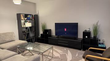 Ein modernes Wohnzimmer mit Flachbildfernseher, Glastisch, Sofa, Sessel und Bücherregal.
