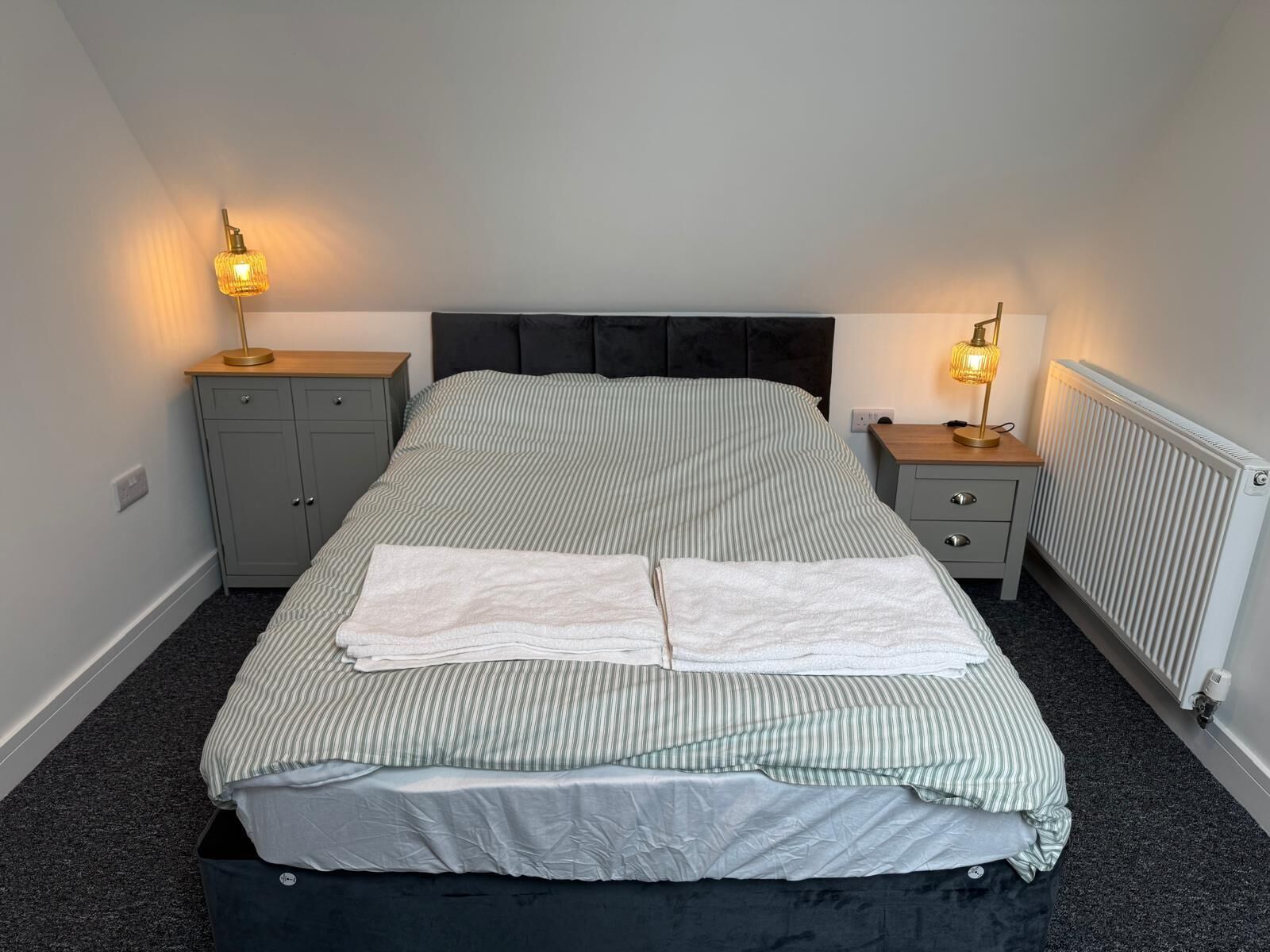 Una cama bien hecha con ropa de cama a rayas y dos lámparas de noche, un armario gris y un radiador.
