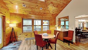 A wooden cabin interior with a dining area, kitchen, and large windows.