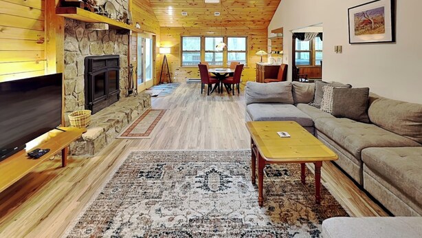 A cozy cabin interior with a stone fireplace, wooden walls, a sofa, a TV, and a dining area.
