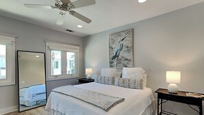 A bedroom with a large bed, bedside lamps, a ceiling fan, a mirror, and a window with natural light. - Tybee Straits 17 (Tybee Island)