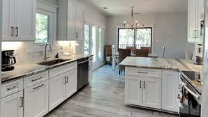 A modern kitchen with white cabinetry, a marble island, and a dining area with wicker chairs. - Tybee Straits 17 (Tybee Island)