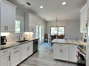A modern kitchen with white cabinetry, a marble island, and a dining area with wicker chairs. - Tybee Straits 17 (Tybee Island)