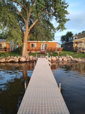 A wooden dock leading to a house with a large tree in the background.