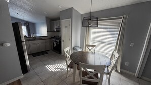A modern kitchen with stainless steel appliances, a dining area with a wooden table and white chairs, and a large window with blinds. - Beach Bums Villa (Davenport)