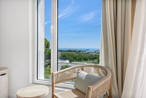 Una silla de mimbre con un cojín junto a una ventana que ofrece vistas a un paisaje y al mar.