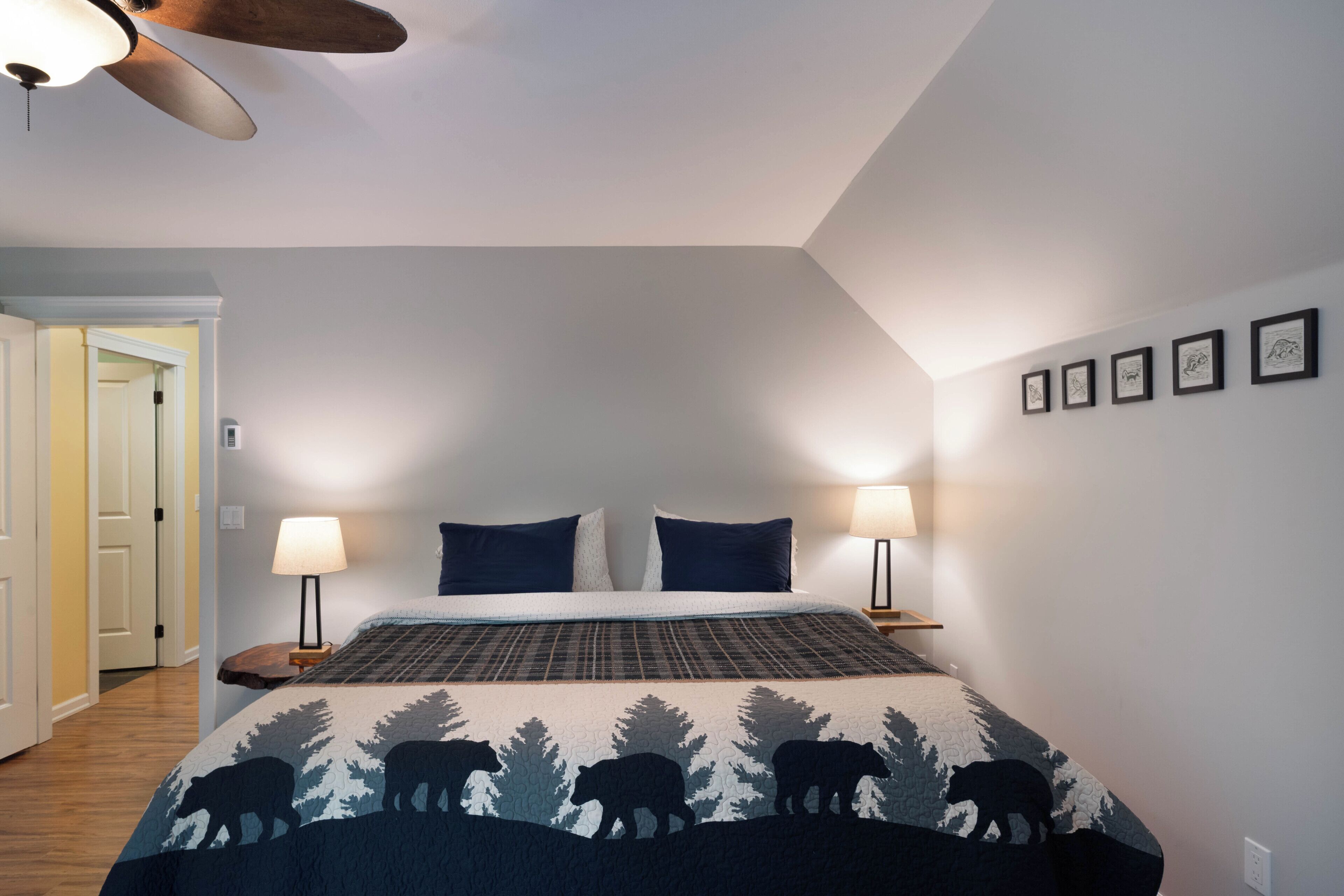 A bedroom with a bed, two bedside lamps, a ceiling fan, and a door leading to another room.