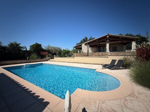 Ein Schwimmbad mit einer Steindeck, einer überdachten Terrasse und Blick auf ein Haus mit Steinverkleidung.
