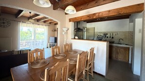A dining area with a wooden table and chairs, a kitchen in the background, and a view of the outdoors. - 3-bedroom family house, swimming pool, garden and terrace near Uzès (Serviers-et-Labaume)