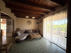 Ein gemütliches Wohnzimmer mit Holzbalkendecke, einer Couch, einem Couchtisch und einem großen Fenster mit transparenten Vorhängen.