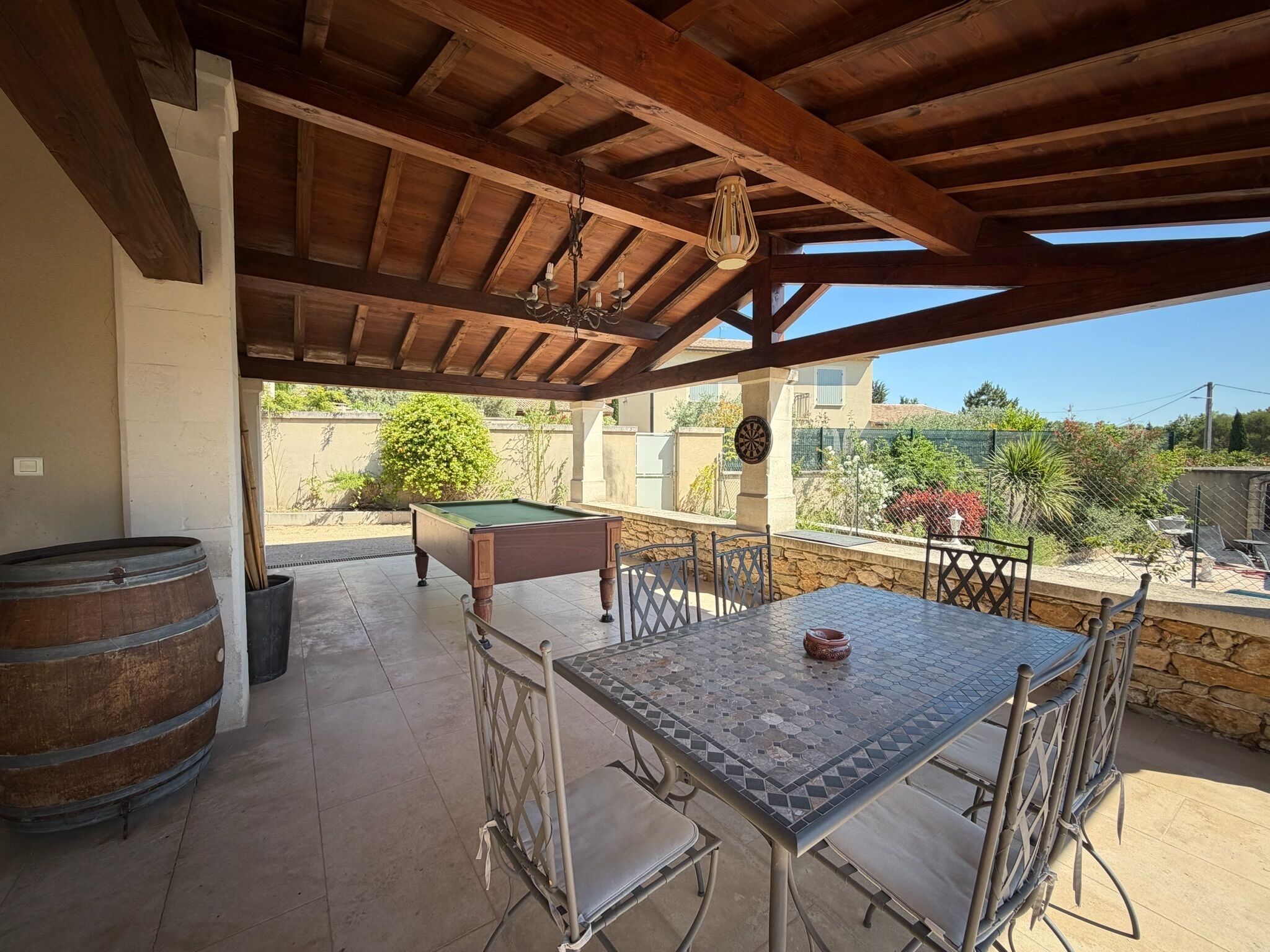 Une terrasse extérieure avec une table de billard, des chaises en métal, une table ornée d’un vase et un tonneau en bois.