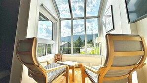 Et værelse med et stort ovenlys, to træstole, et lille bord og udsigt til et hus og træer.