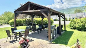 A covered outdoor seating area with tables and chairs, surrounded by greenery and a view of a residential area.