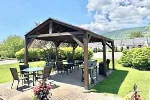 A covered outdoor seating area with tables and chairs, surrounded by greenery and a view of a residential area.