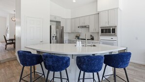 A modern kitchen with white cabinetry, a central island with bar stools, stainless steel appliances, and a dining area with a wooden table and chairs. - Greyhawk Golf Escape w/ 5-Bedrooms, Heated Pool, Spa & Putting Green (Scottsdale)