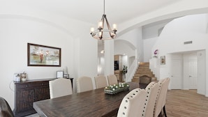 A dining room with a large wooden table, white chairs, a chandelier, and a staircase in the background. - Greyhawk Golf Escape w/ 5-Bedrooms, Heated Pool, Spa & Putting Green (Scottsdale)