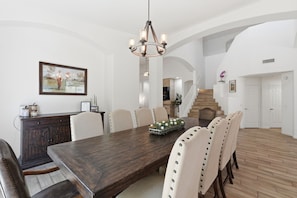 Ein Esszimmer mit einem großen Holztisch, weißen Stühlen, einer Chandelierleuchtre und einer Treppe im Hintergrund.
