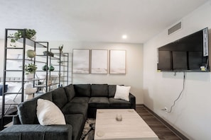 Ein modernes Wohnzimmer mit einem Ecksofa, einem Couchtisch, einem Pflanzenregal und einem Wandfernseher.
