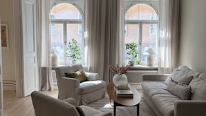 Ett rymligt vardagsrum med en soffa, en fåtölj och ett soffbord, stora fönster med gardiner och utsikt över omgivningen.