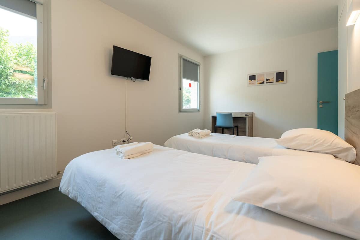 Habitación de hotel con dos camas, un televisor montado en la pared, un escritorio con una silla y una ventana con vistas a zonas verdes.
