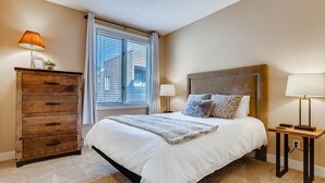 A bedroom with a bed, a dresser, a nightstand, and a window with blinds. - Sun Vail 21b, 3BD Condo With Vail Mountain Views (Vail)