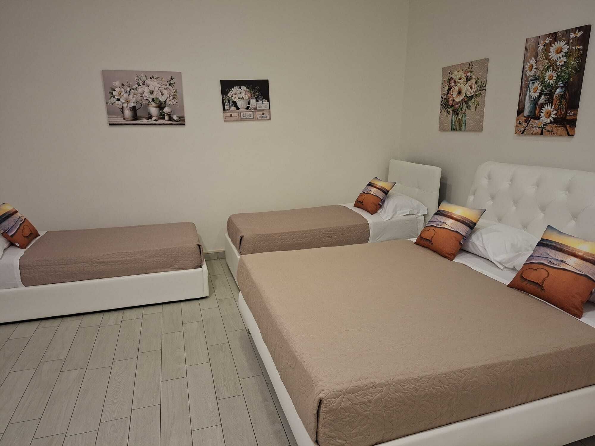 A hotel room with two single beds, a larger bed, and a white headboard. There are three wall-mounted artworks depicting floral arrangements.