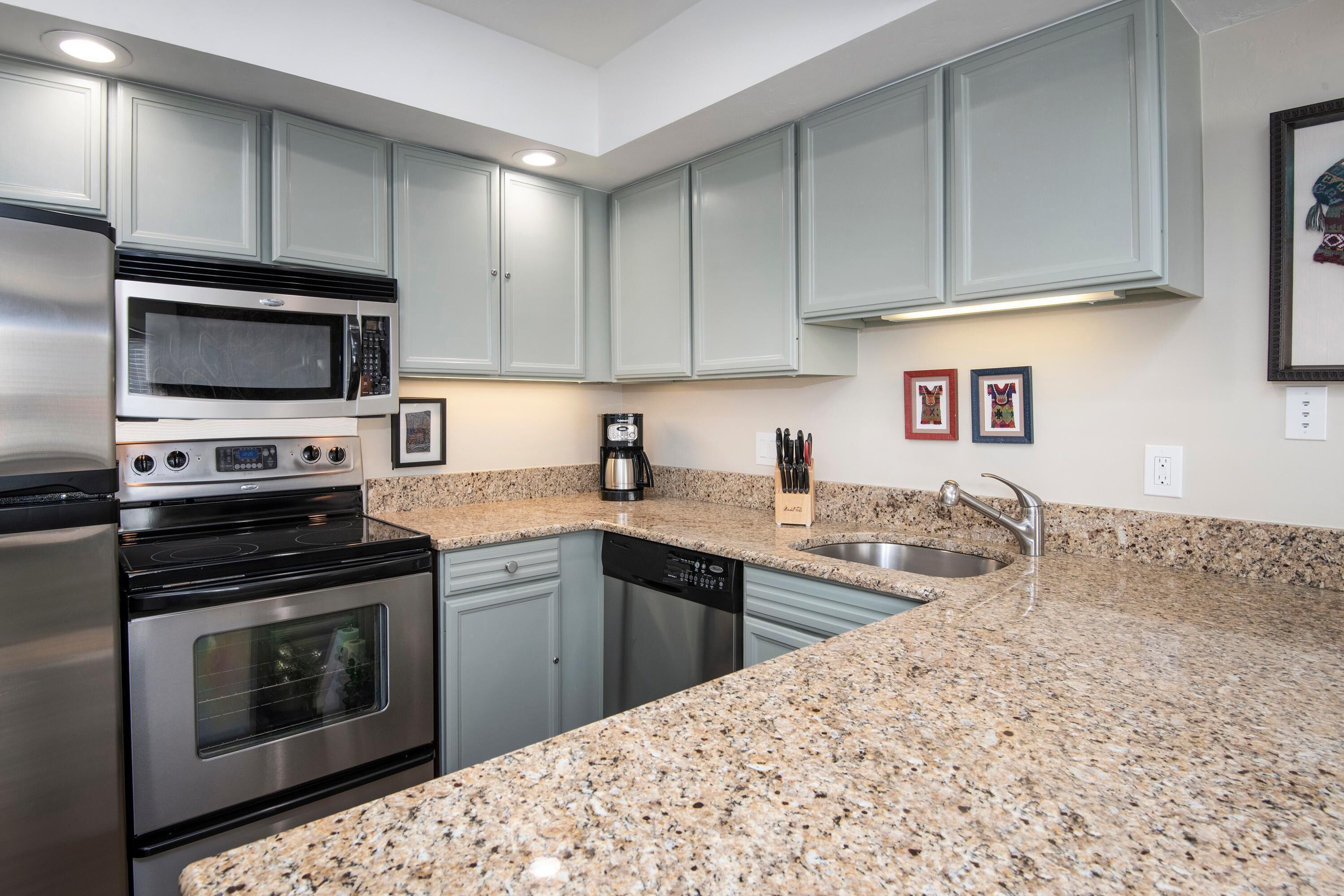 Una cocina con microondas de acero inoxidable, horno y lavavajillas, encimeras de granito y muebles de cocina color gris claro.