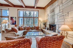 A cozy living room with a stone fireplace, large windows, and a comfortable sofa. - Treetops 5C, 3BD Ski-in Ski-out Condo (Vail)