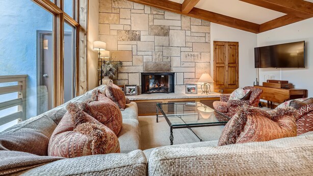 A cozy living room with a stone fireplace, wooden beams, a sofa set, and a flat-screen TV. - Treetops 5C, 3BD Ski-in Ski-out Condo (Vail)