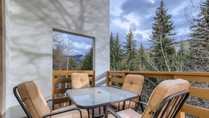 A balcony with a glass table and four cushioned chairs, overlooking a forested area with mountains in the background. - Treetops 5C, 3BD Ski-in Ski-out Condo (Vail)