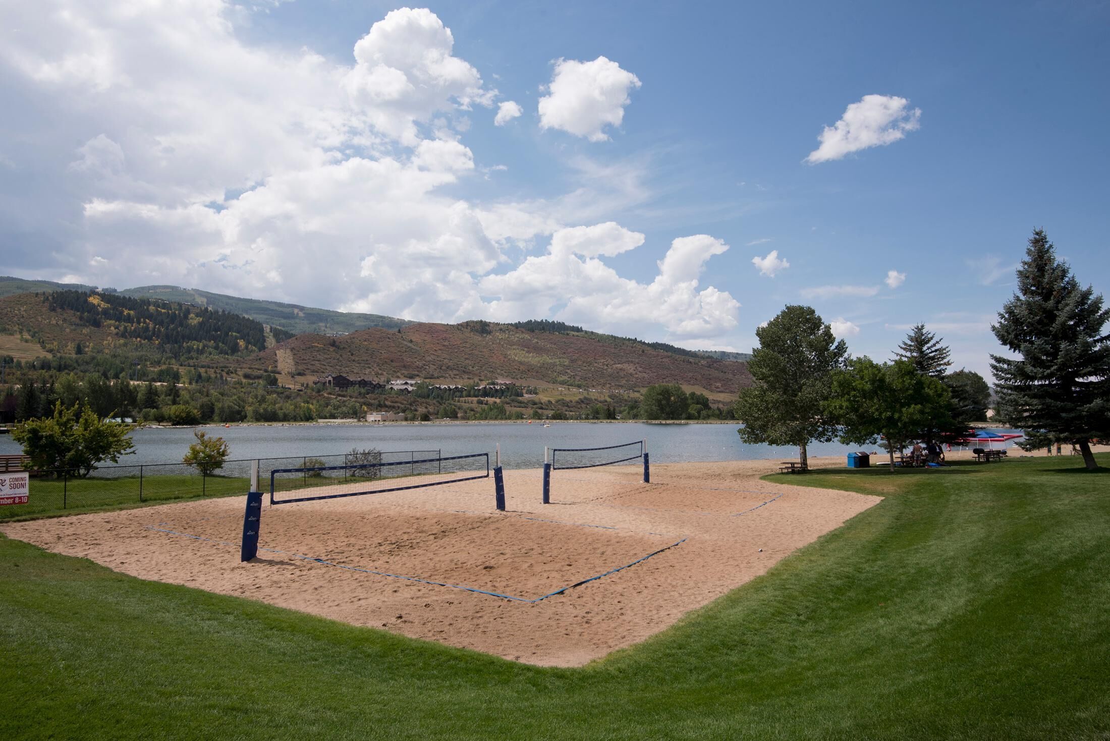 Cancha de voleibol de playa con red, rodeada de césped y árboles, con vista a un lago y colinas al fondo.