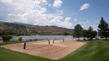 Cancha de voleibol de playa con red, rodeada de césped y árboles, con vista a un lago y colinas al fondo.