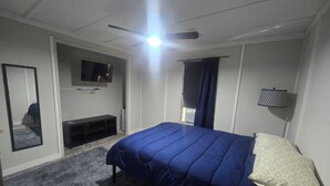 Una camera da letto con un letto, una TV, una ventola a soffitto, una finestra con tende e una lampada da terra.