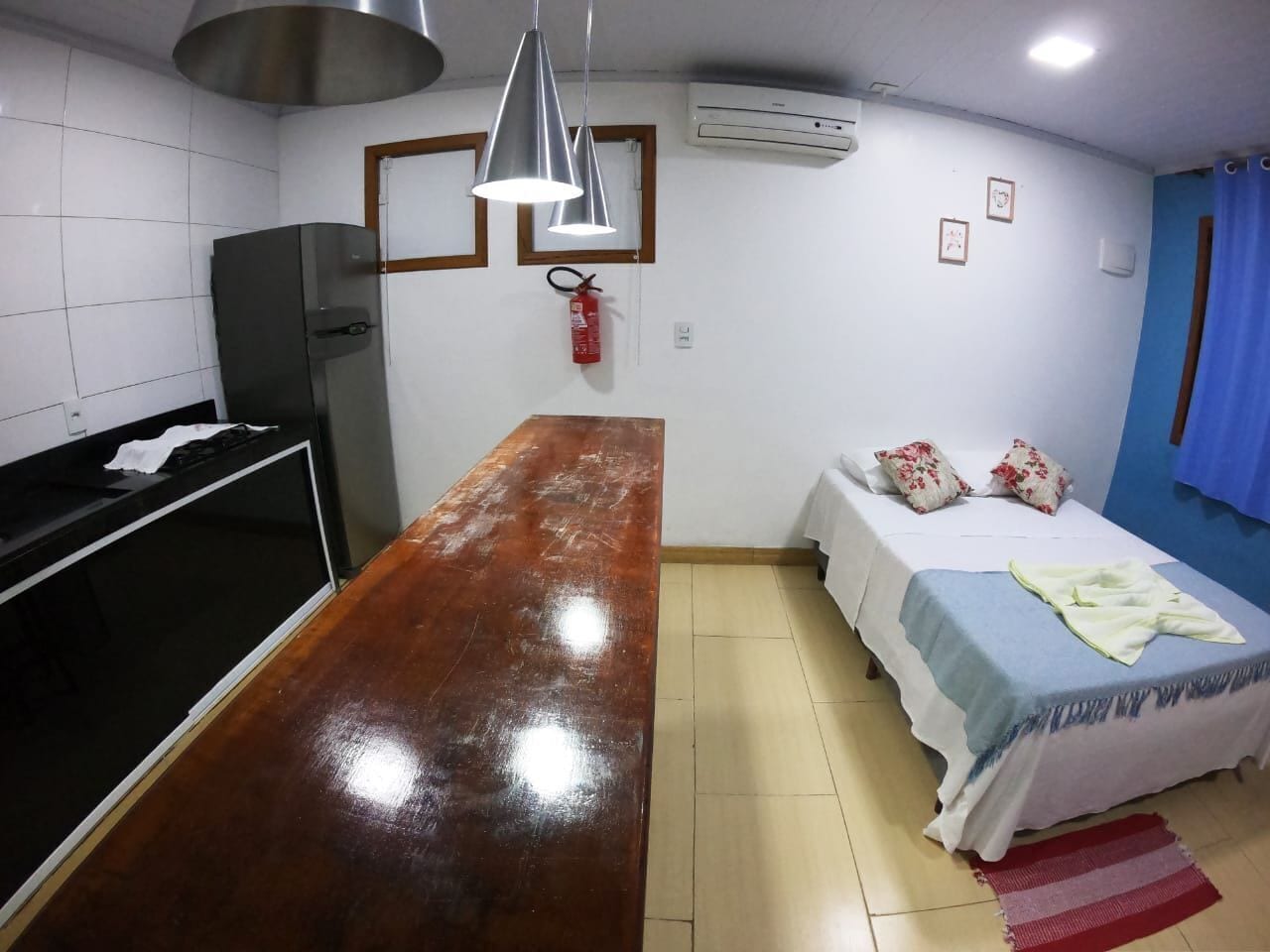A compact hotel room with a kitchenette, a bed with floral pillows, a blue curtain, and a red fire extinguisher.