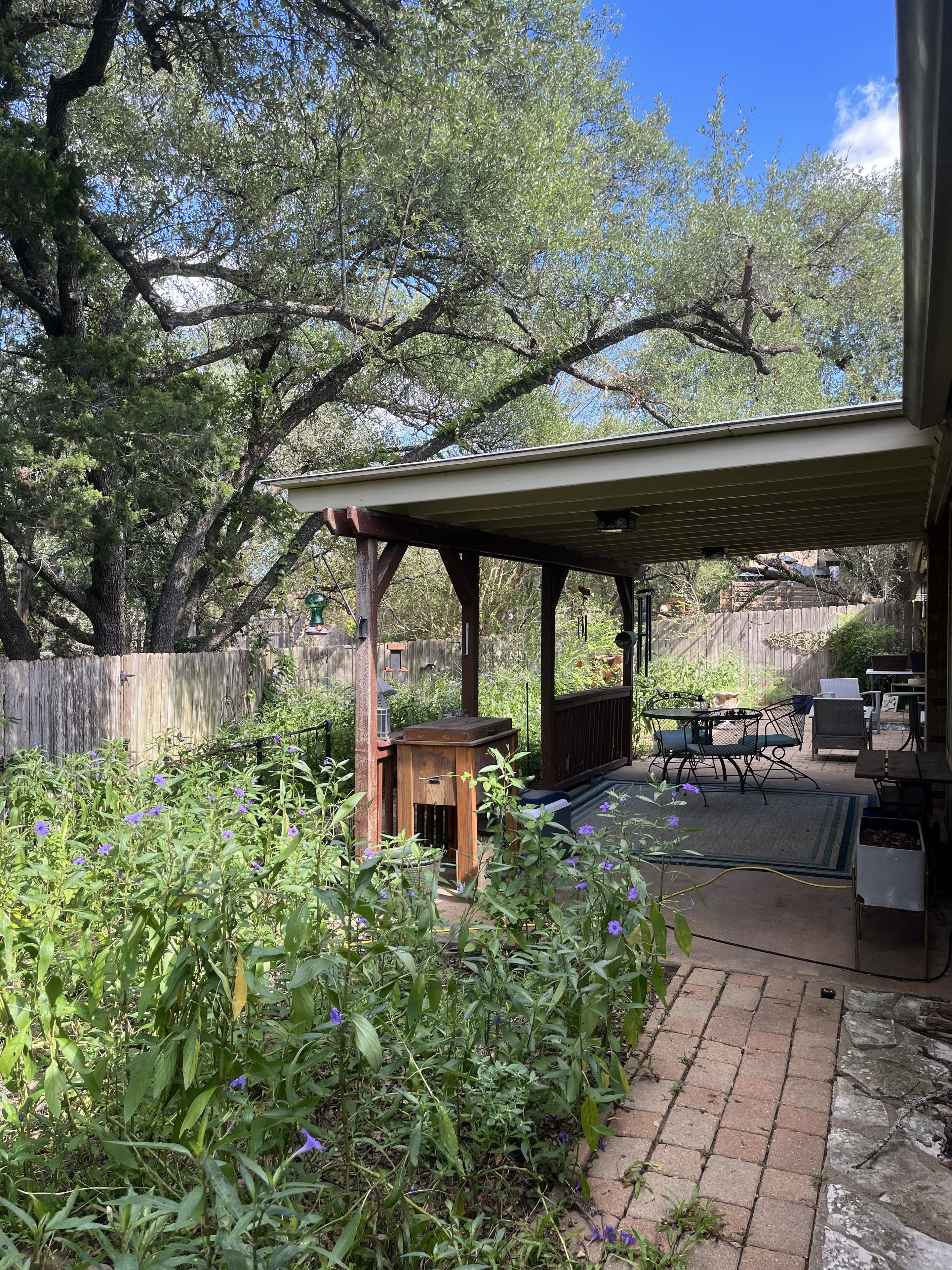 A covered patio area with a wooden table and chairs, surrounded by greenery and a wooden fence.