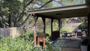 A covered patio area with a wooden table and chairs, surrounded by greenery and a wooden fence.