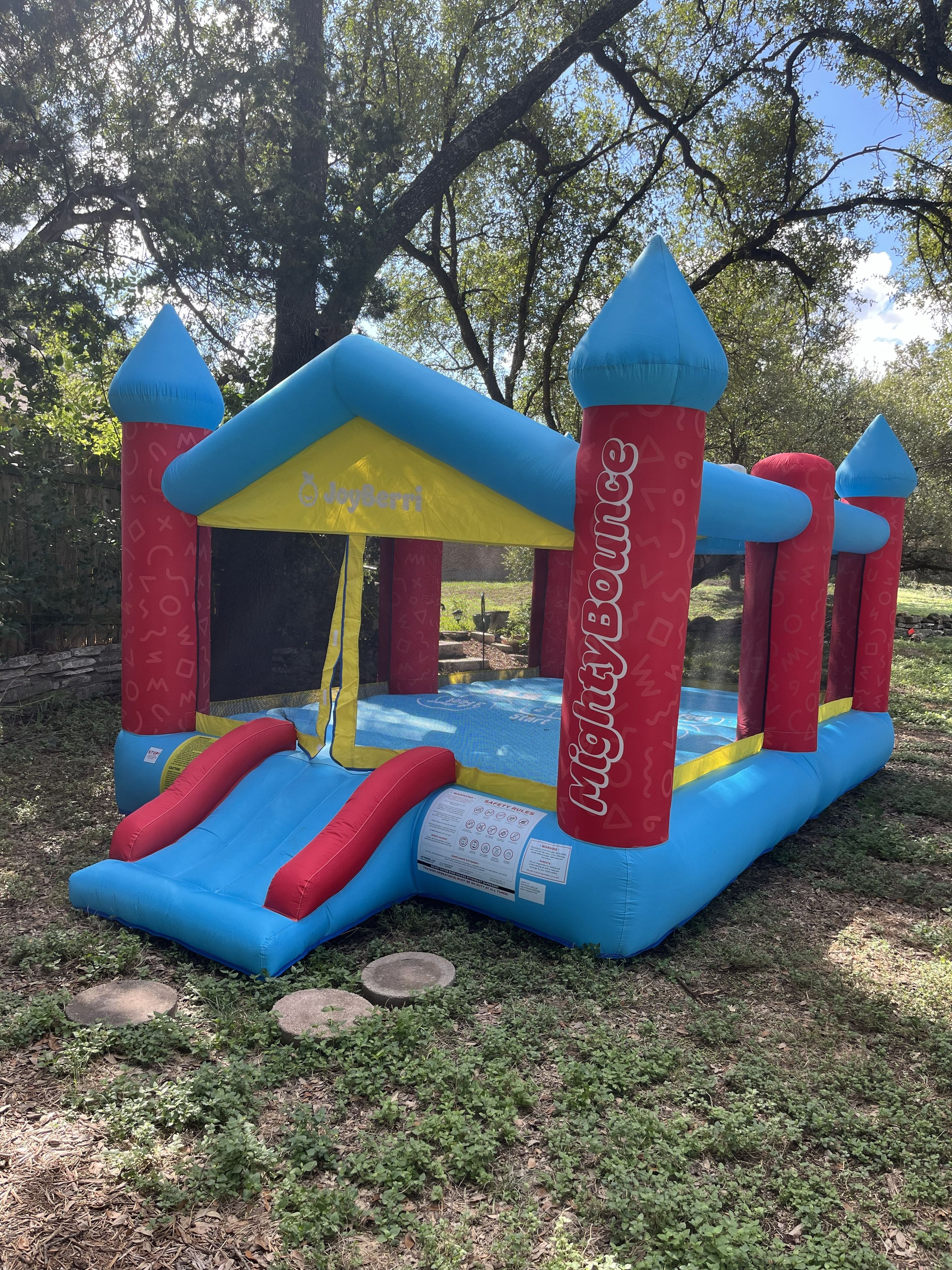 An inflatable bounce house with a slide, labeled 'JayBerri' and 'Light Bounce,' set in a grassy area with trees in the background.