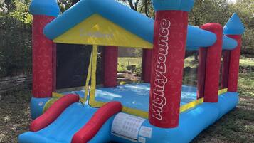 An inflatable bounce house with a slide, labeled 'JayBerri' and 'Light Bounce,' set in a grassy area with trees in the background.
