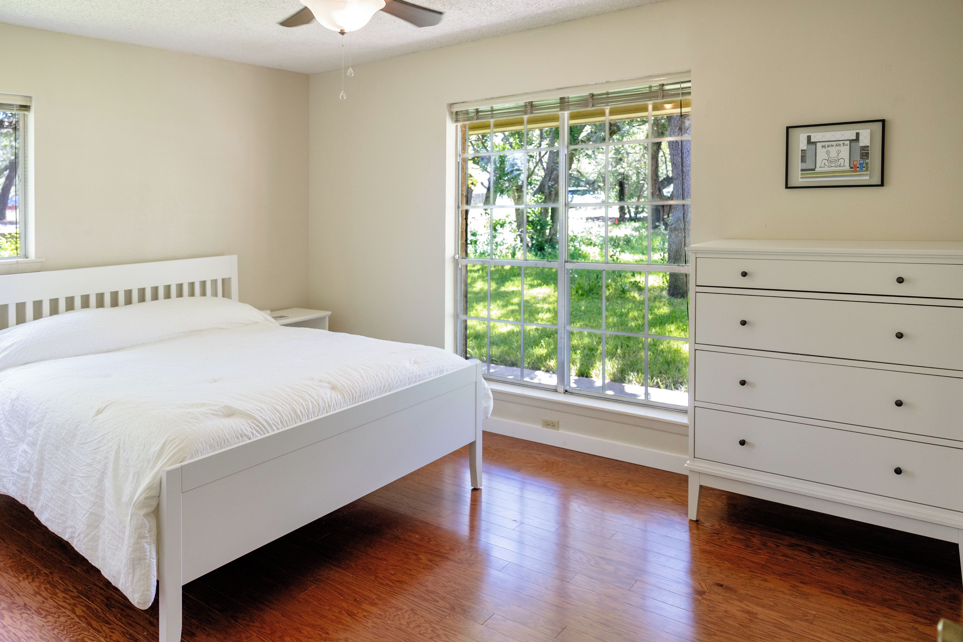 A bedroom with a bed, a dresser, a window with a view of greenery, and a ceiling fan.