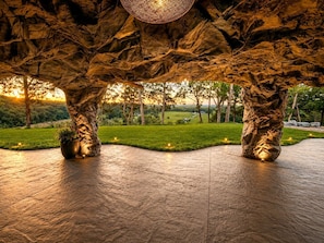 A stone-tiled floor with a unique rock-like structure overhead, a potted plant, and a view of a green landscape.