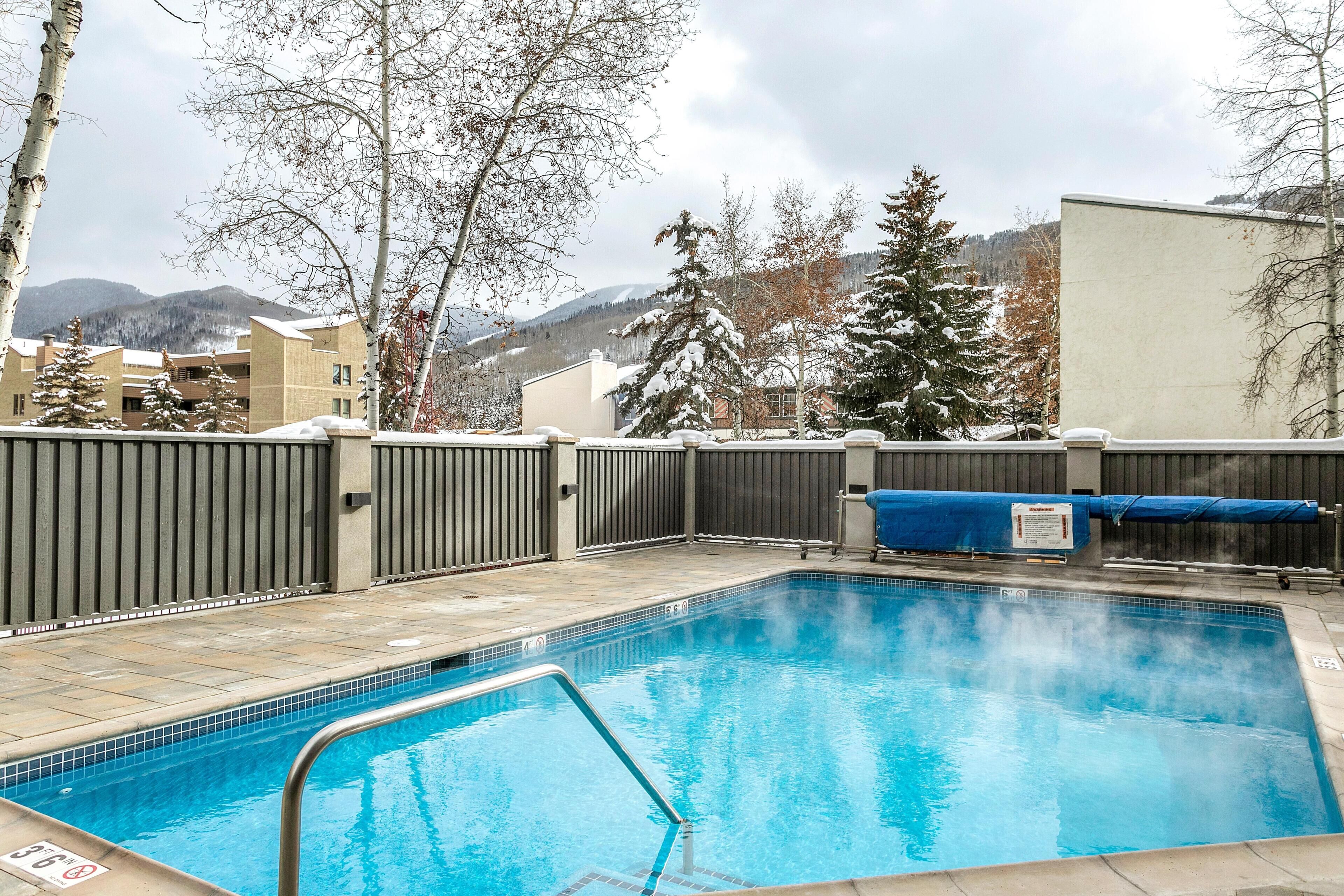 A swimming pool with a metal ladder, surrounded by a fence, with a view of mountains and trees in the background.