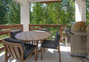 Eine überdachte Terrasse mit einem runden Tisch, vier Stühlen, einem Grill und Blick auf Bäume.