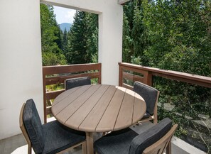 Ein runder Holztisch mit vier Stühlen, eine weiße Wand und Blick auf einen Wald und ein Gebirge.